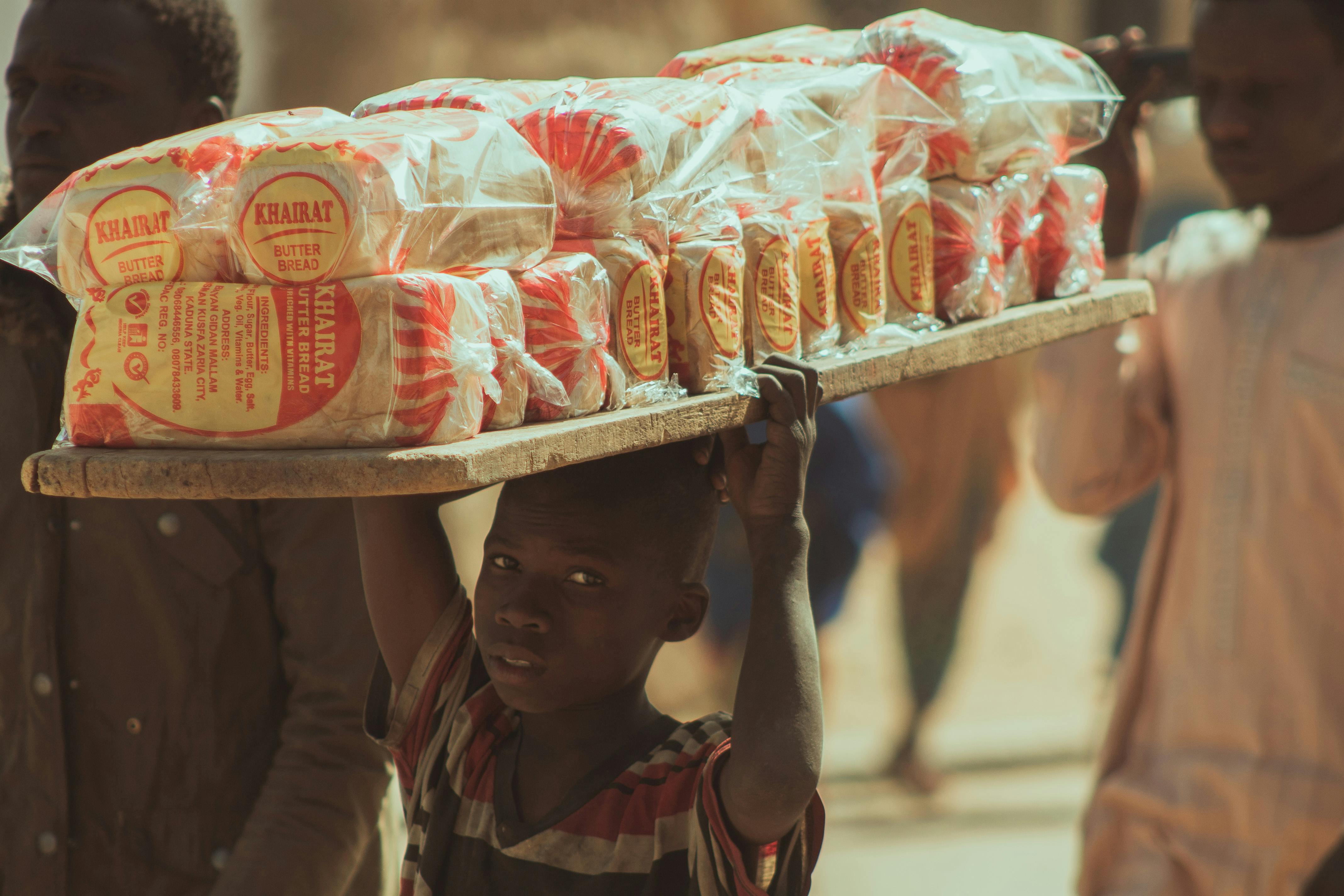 A young boy carries packaged bread on his head outdoors, highlighting daily life and resilience.