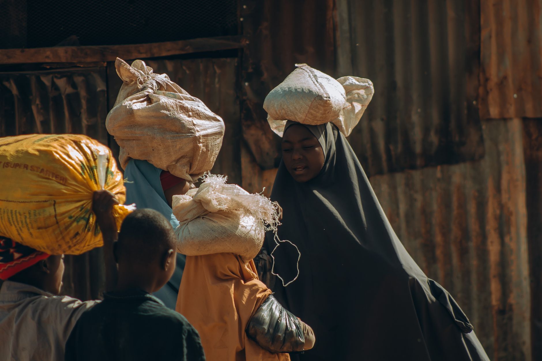 Women in traditional attire carry goods at an outdoor market in vibrant daylight.