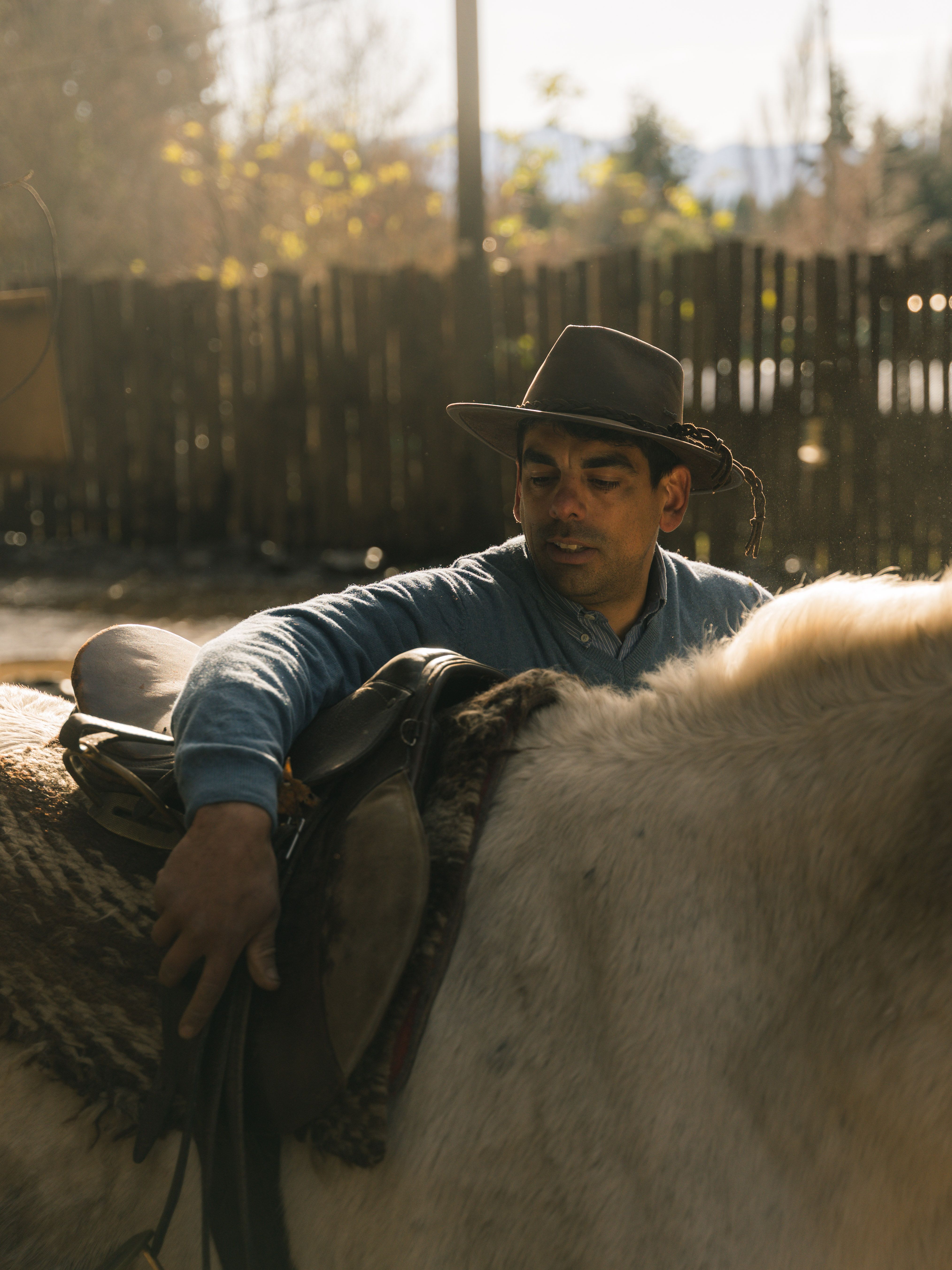 A man wearing a hat saddling a horse outdoors near a wooden fence in natural light.