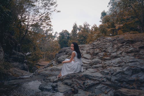 Free Woman Wearing White Dress Sitting on Rock Near Trees Stock Photo