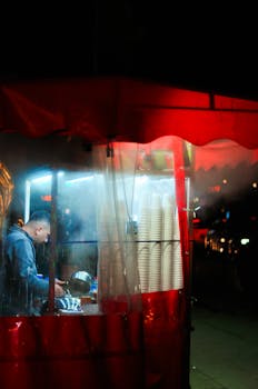 A street food vendor inside a red tent preparing food at night, illuminated by bright lights.
