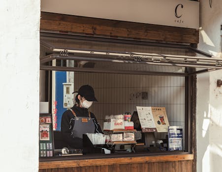 Photo by 女子 正真 A worker behind the counter of an outdoor cafe kiosk, preparing orders.