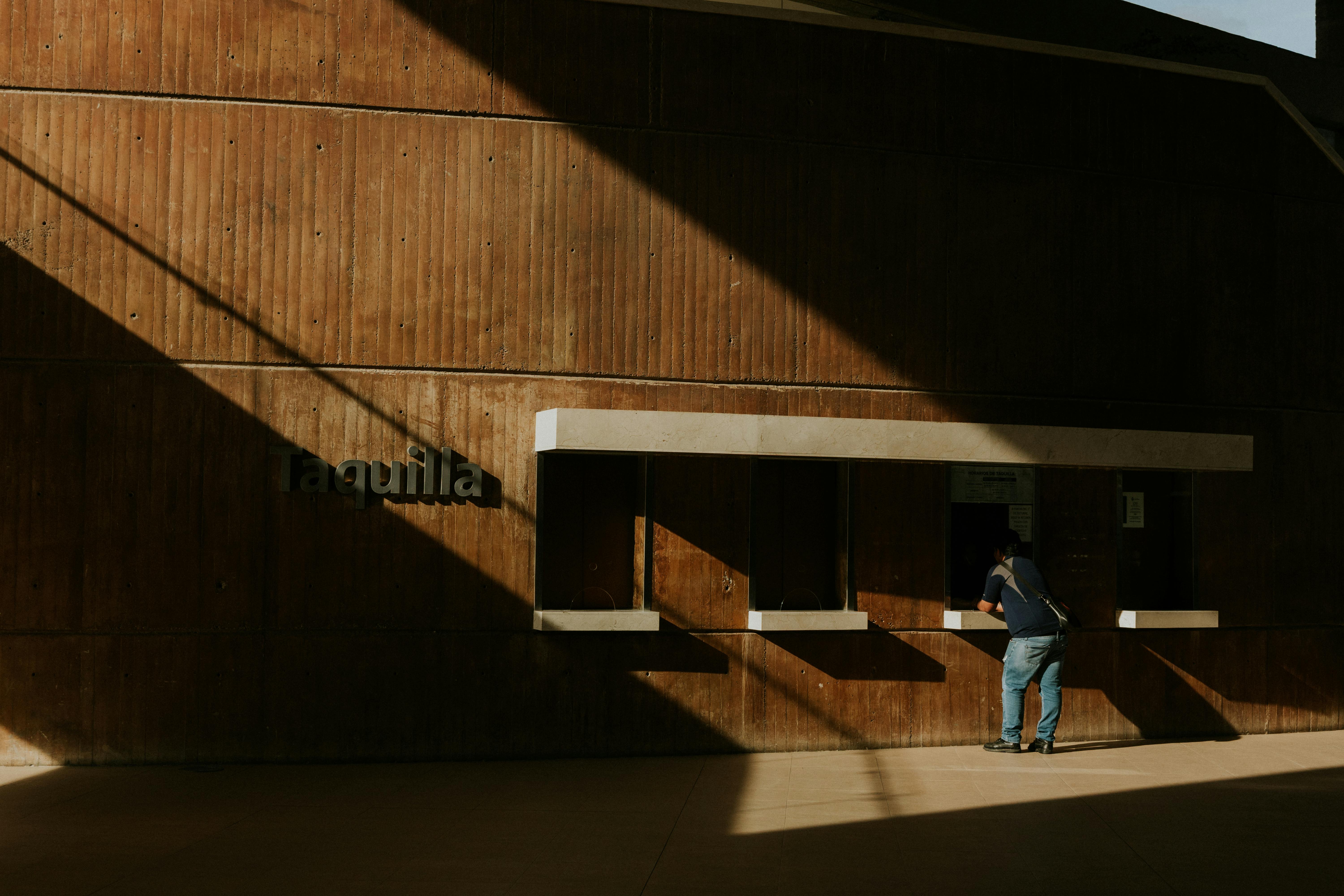 A person leans into a sunlit ticket booth, casting dramatic shadows on a textured wall.