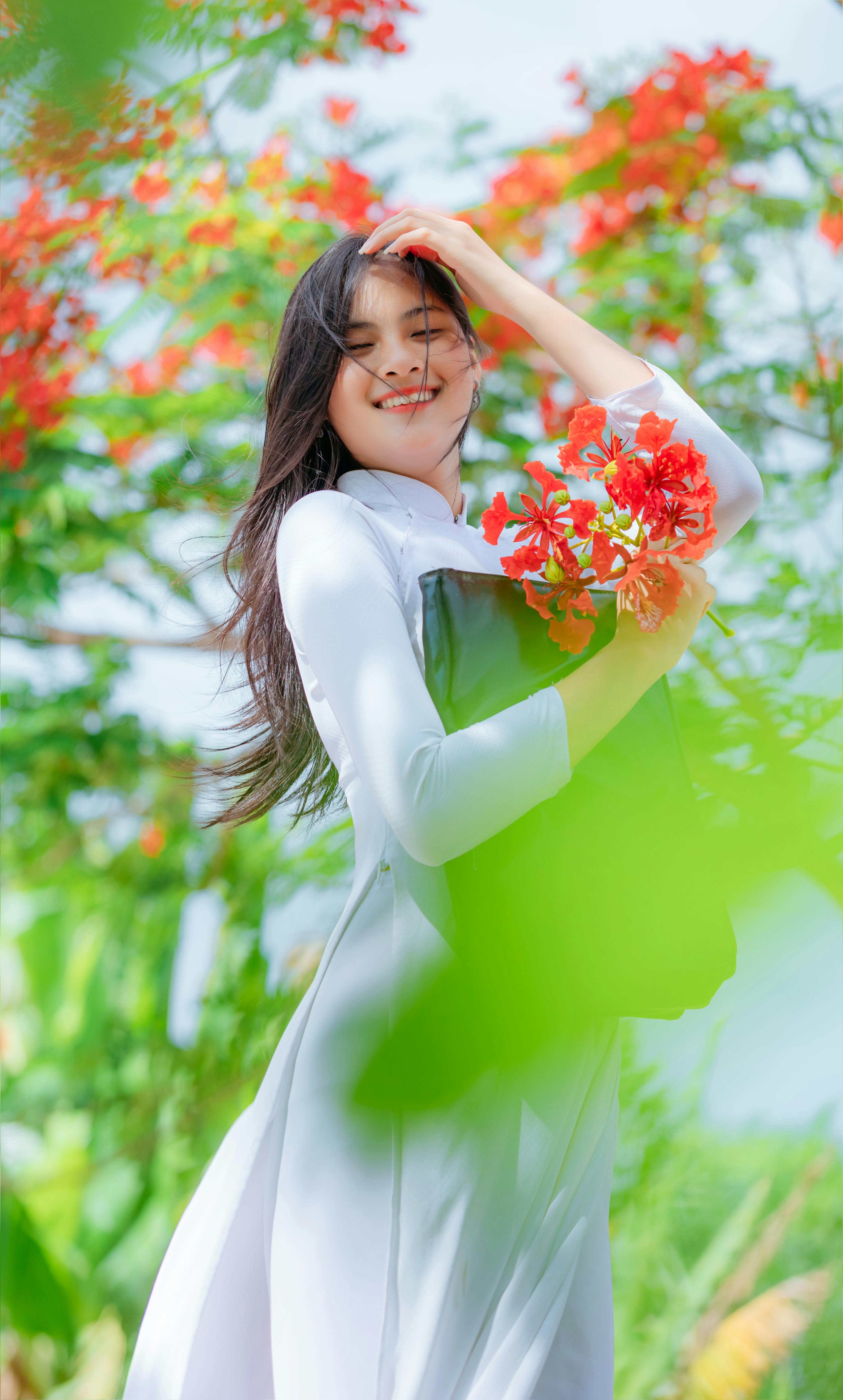 A cheerful woman stands outdoors in a white dress, holding vibrant red flowers under a bright summer sky.