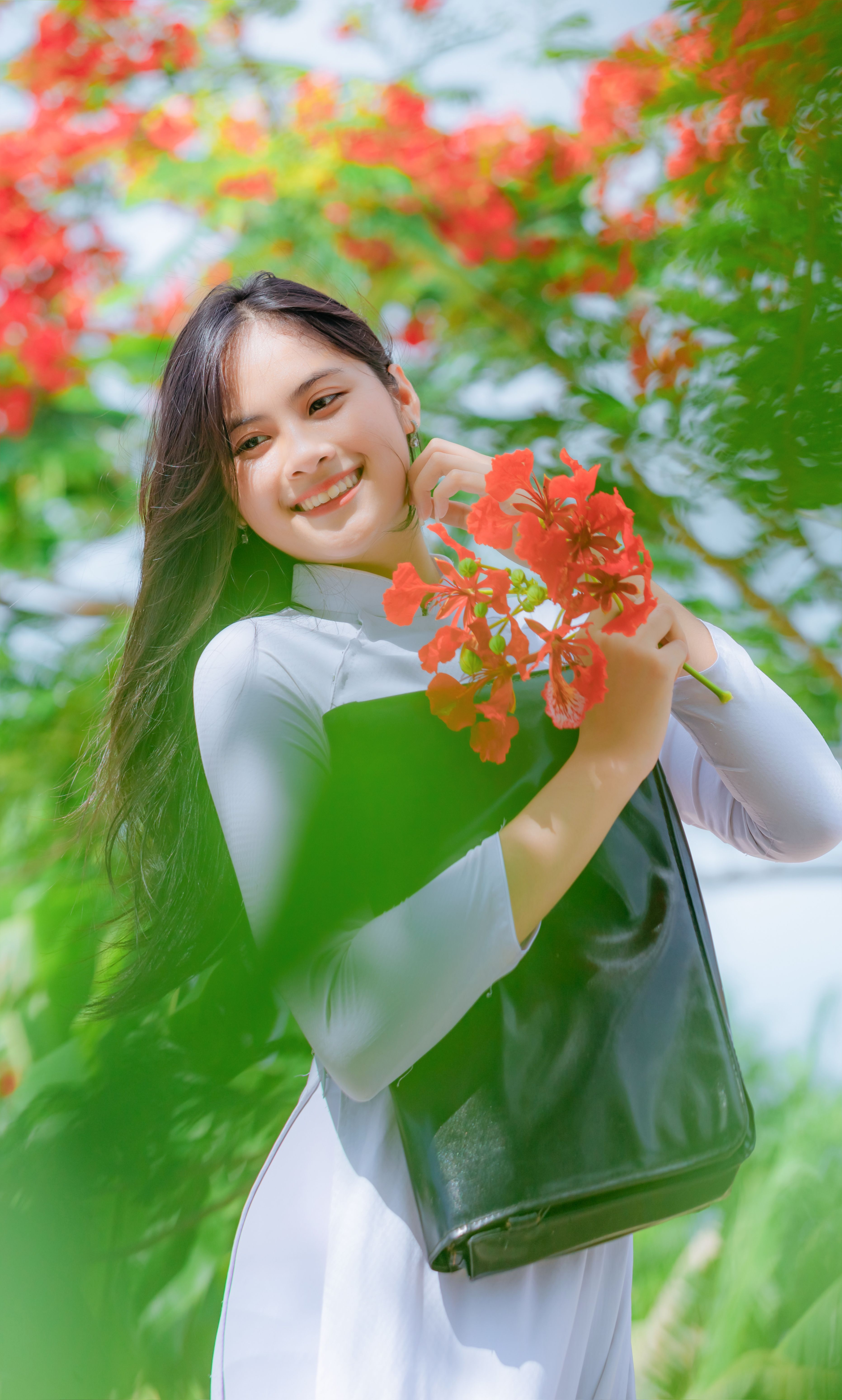 Smiling woman in a traditional white Ao Dai holding vibrant red flowers, outdoors in natural light.