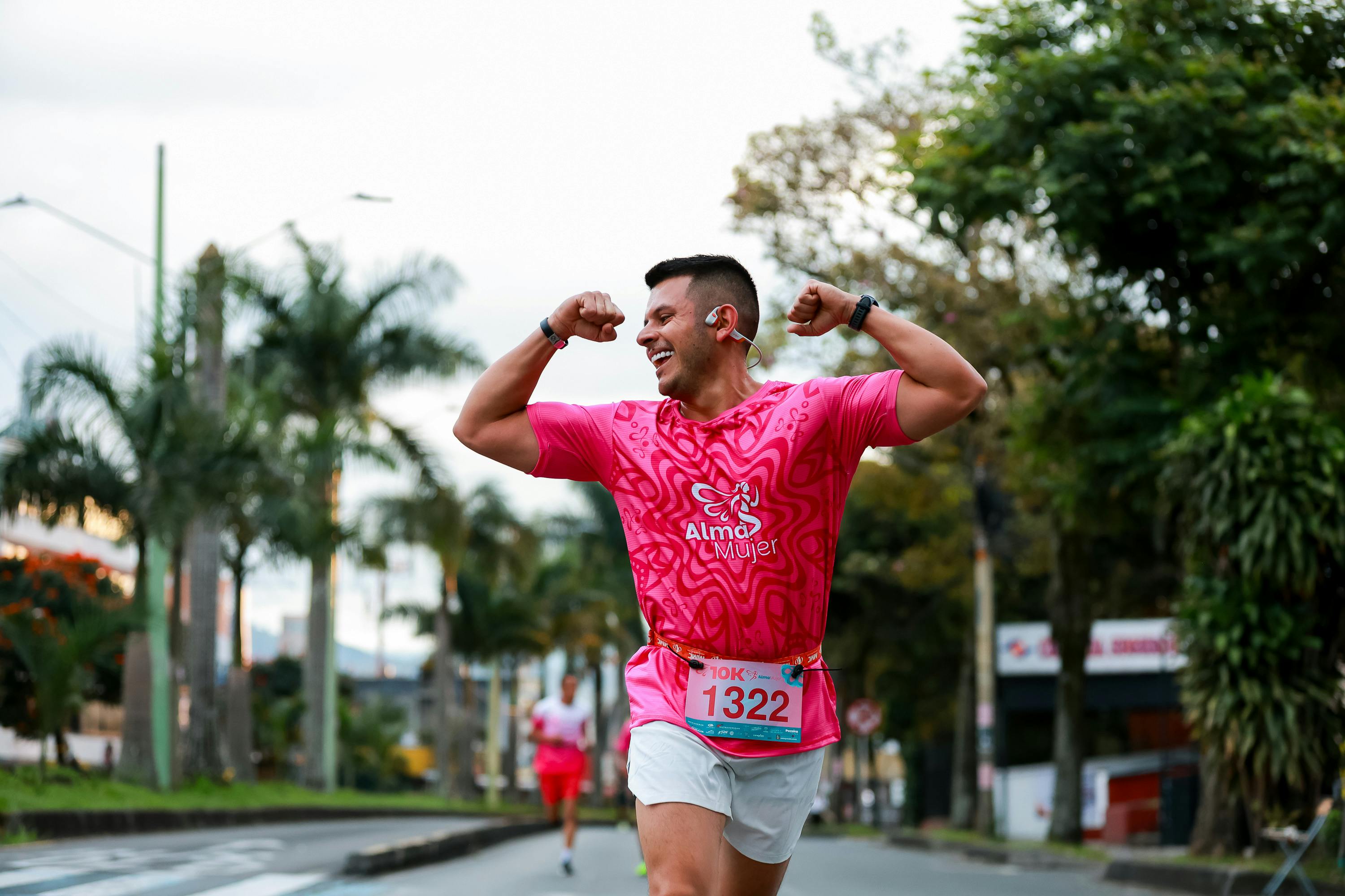 Smiling athlete in pink shirt flexing muscles while running a race outdoors.
