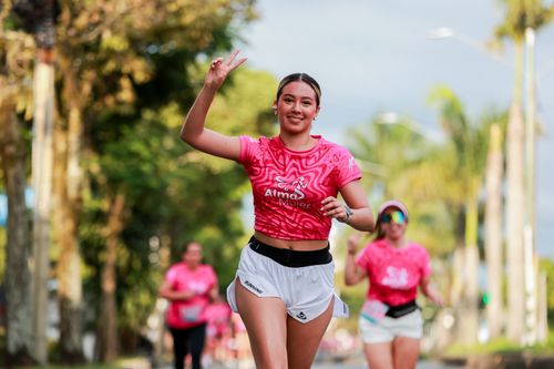 Free Energetic woman running outdoors in pink event shirt, smiling and flashing a peace sign. Stock Photo
