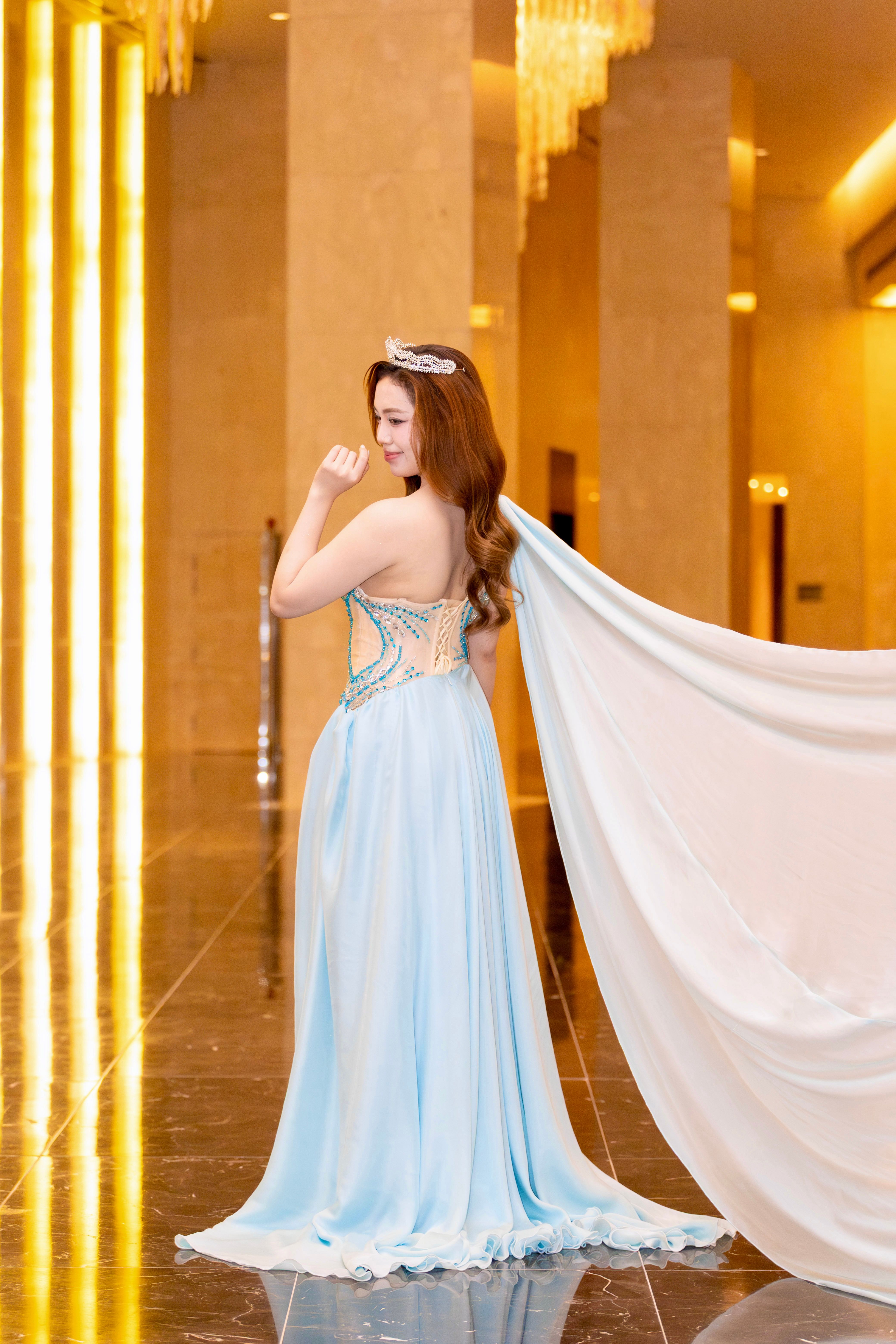 A woman in a blue gown and tiara poses in a lavish indoor setting in Hà Nội, Việt Nam.