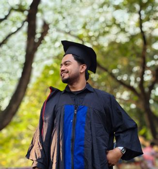 A young adult male in graduation attire smiles confidently in a sunlit park setting.