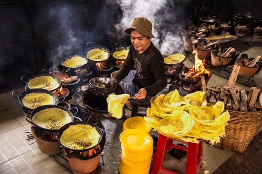 Asian street vendor preparing traditional yellow pancakes over open fires. Vibrant and lively scene.