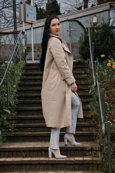 Stylish woman in beige trench coat poses on garden stairs in Bremen, Germany, showcasing modern fashion.