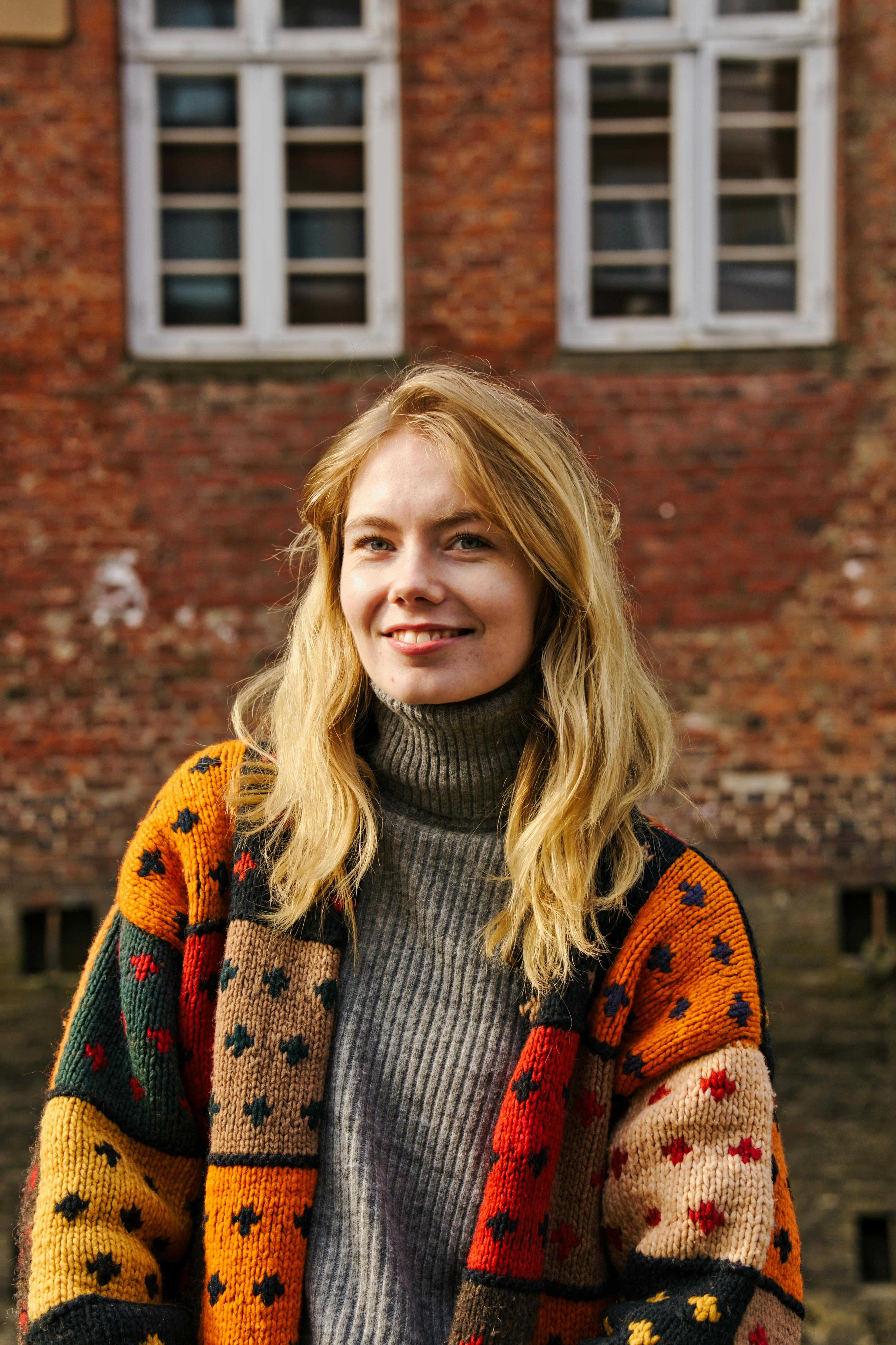 A cheerful young woman in a colorful sweater poses outdoors against a historic brick building.