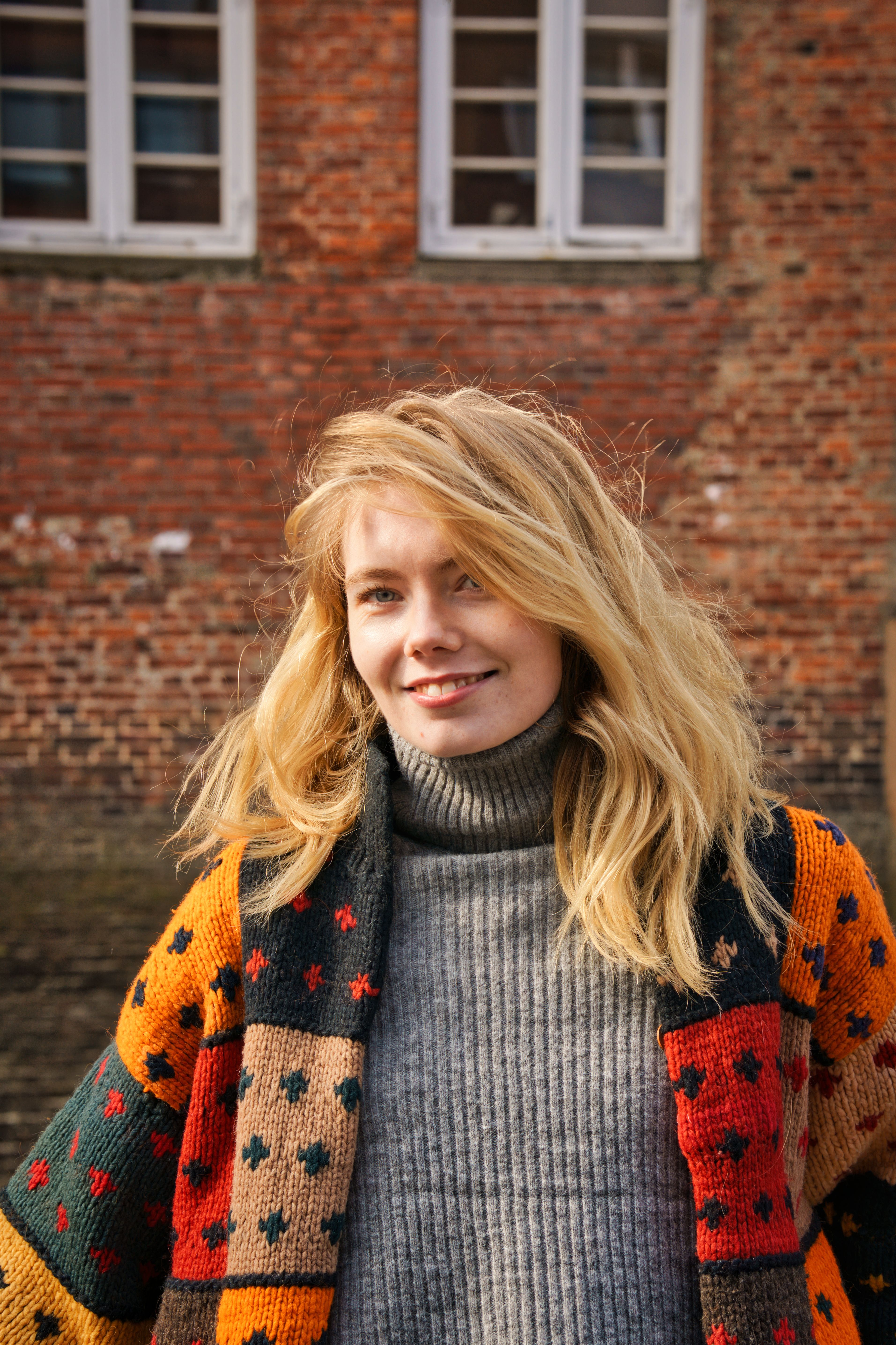 A cheerful woman wearing a colorful sweater smiles brightly in front of a brick wall under daylight.