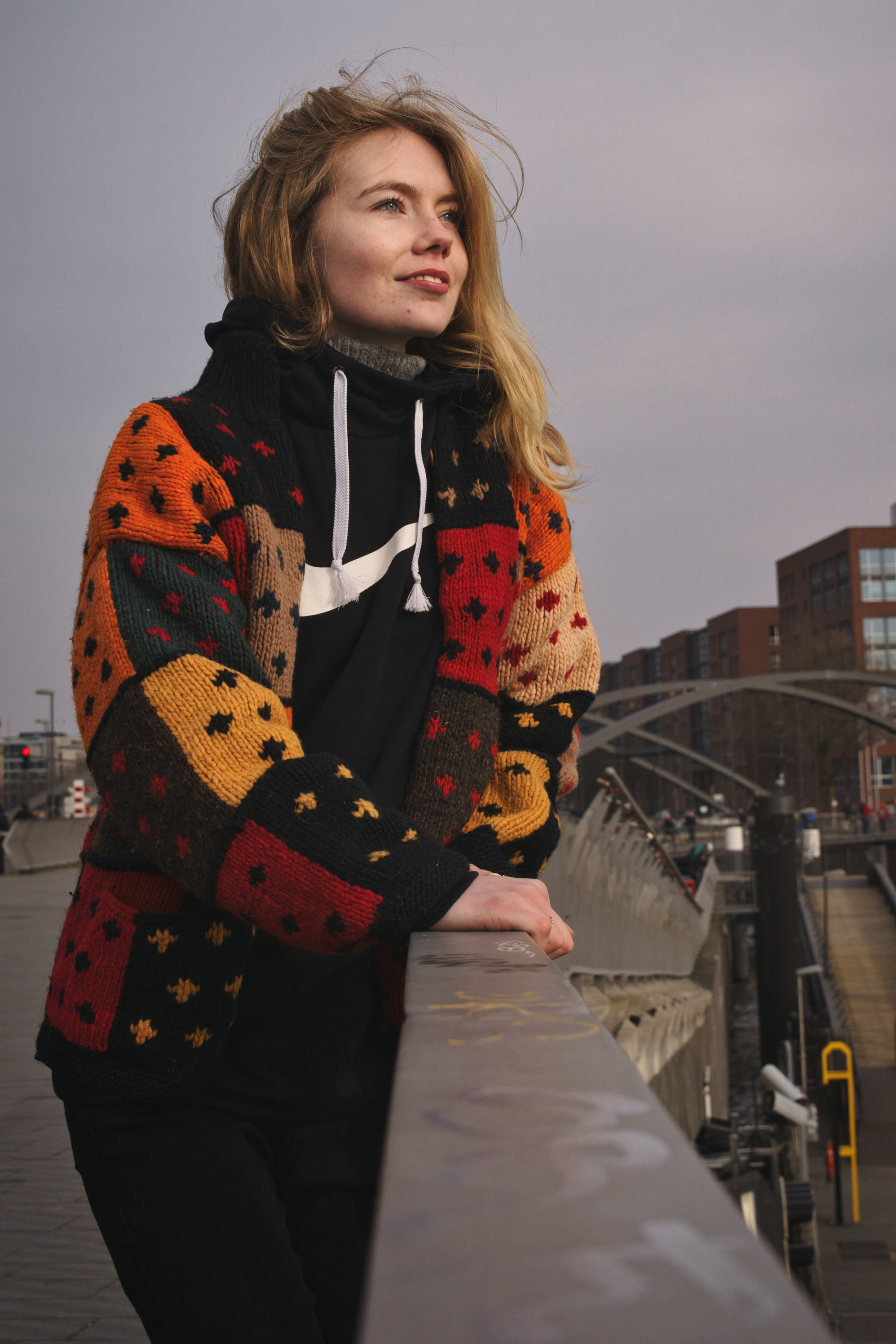 A woman in a colorful knit sweater leans on a railing, with urban Hamburg in the background.