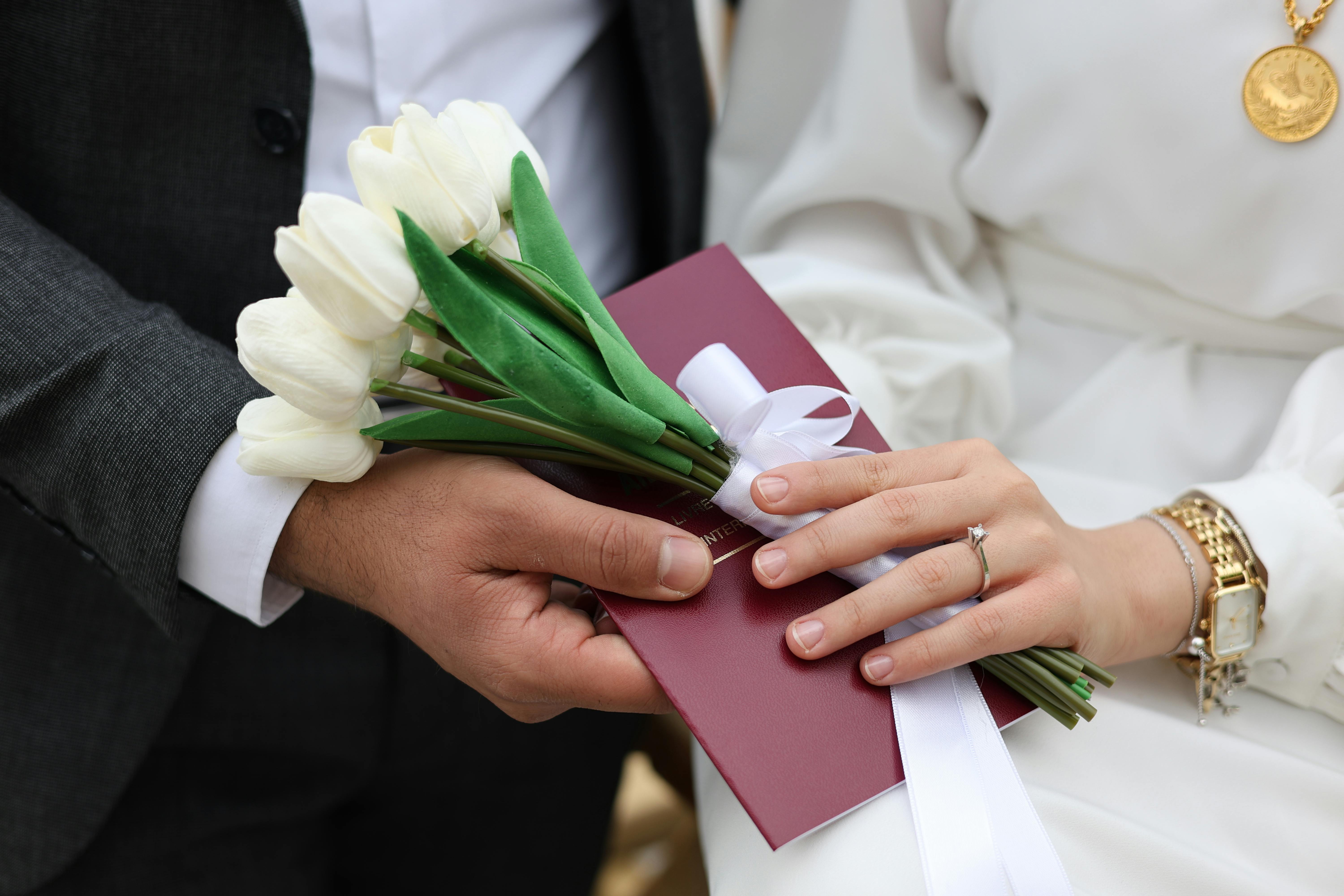 Close-up of a bride and groom holding white tulips and a red envelope during a wedding ceremony.