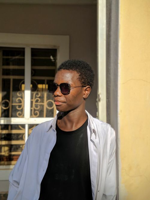 Free Casual portrait of a young man wearing sunglasses standing by a window, with sunlight casting gentle shadows. Stock Photo