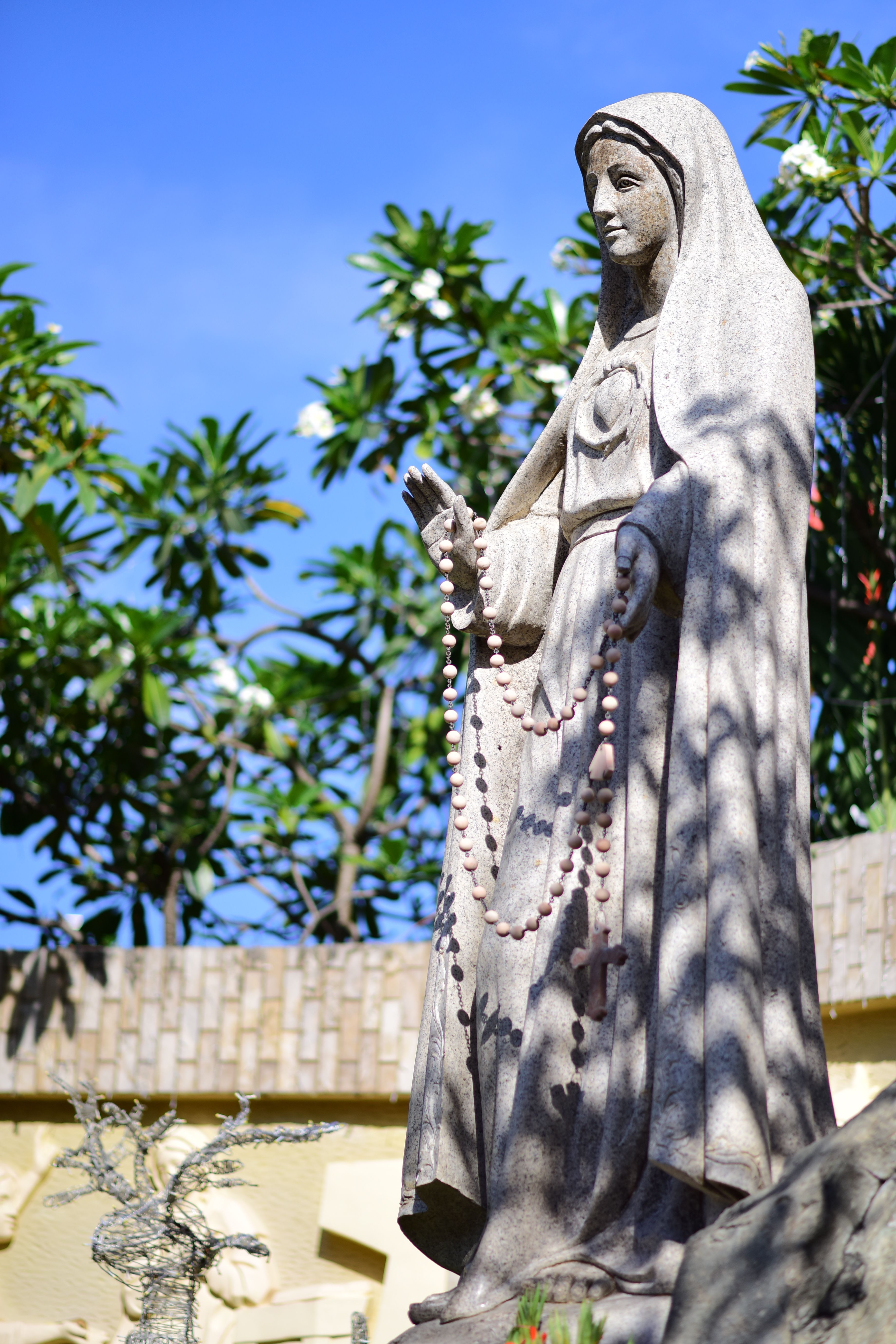 Outdoor statue of Virgin Mary with rosary beads under blue sky and trees.