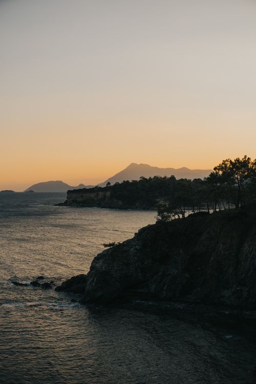 Free Beautiful coastal sunset with cliffs and mountains silhouetted against an orange sky, ocean waves below. Stock Photo