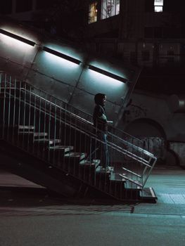 A person stands contemplatively on urban stairs at nighttime, illuminated by cool lights.