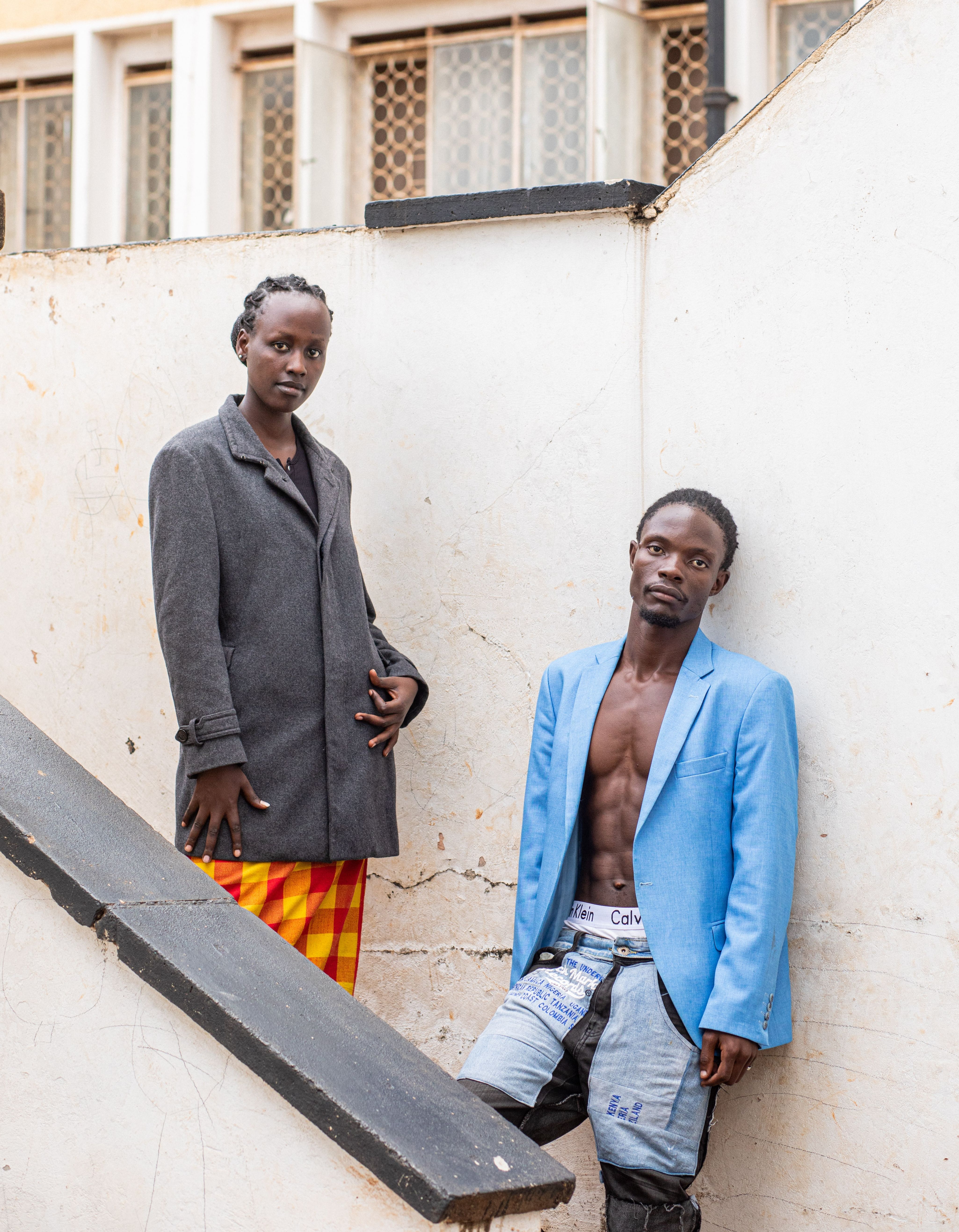 Two individuals in fashionable outfits pose in an urban setting.