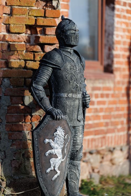 Free Statue of a medieval knight in armor with shield featuring lion emblem, standing against a brick wall. Stock Photo