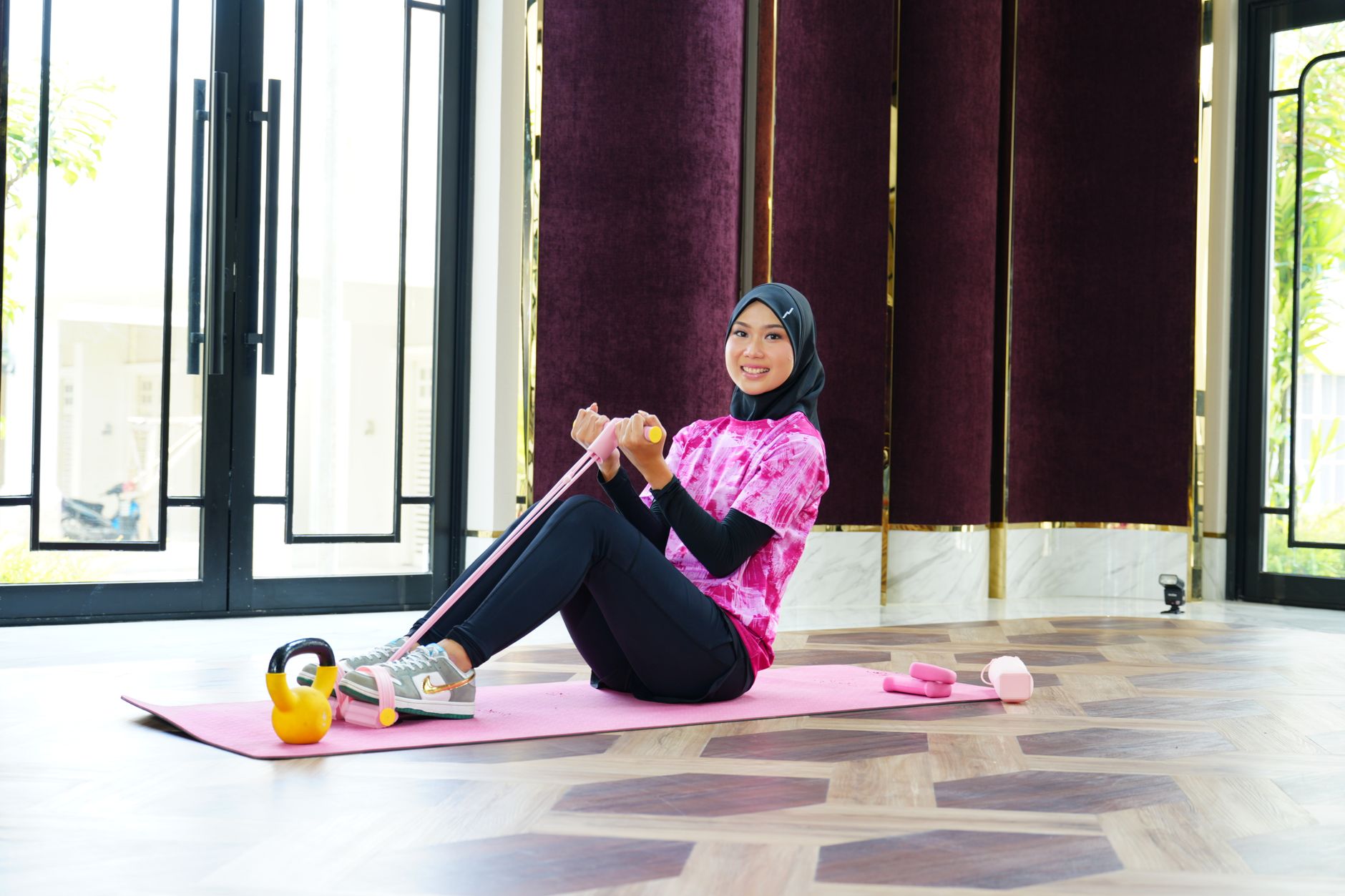 A woman in sporty attire and hijab exercises indoors using a resistance band, showcasing fitness and fashion.