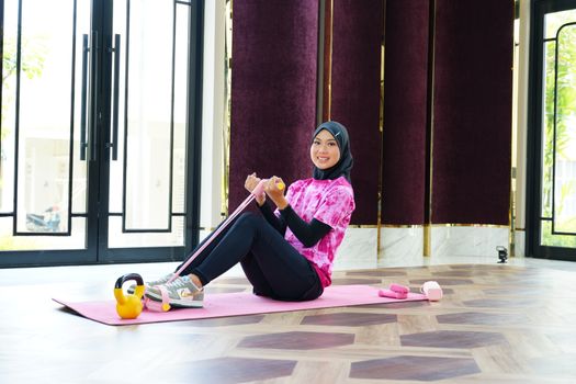 A woman in sporty attire and hijab exercises indoors using a resistance band, showcasing fitness and fashion.