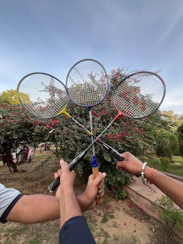 Three badminton rackets raised outdoors against a vibrant flowering bush, symbolizing sport and leisure.