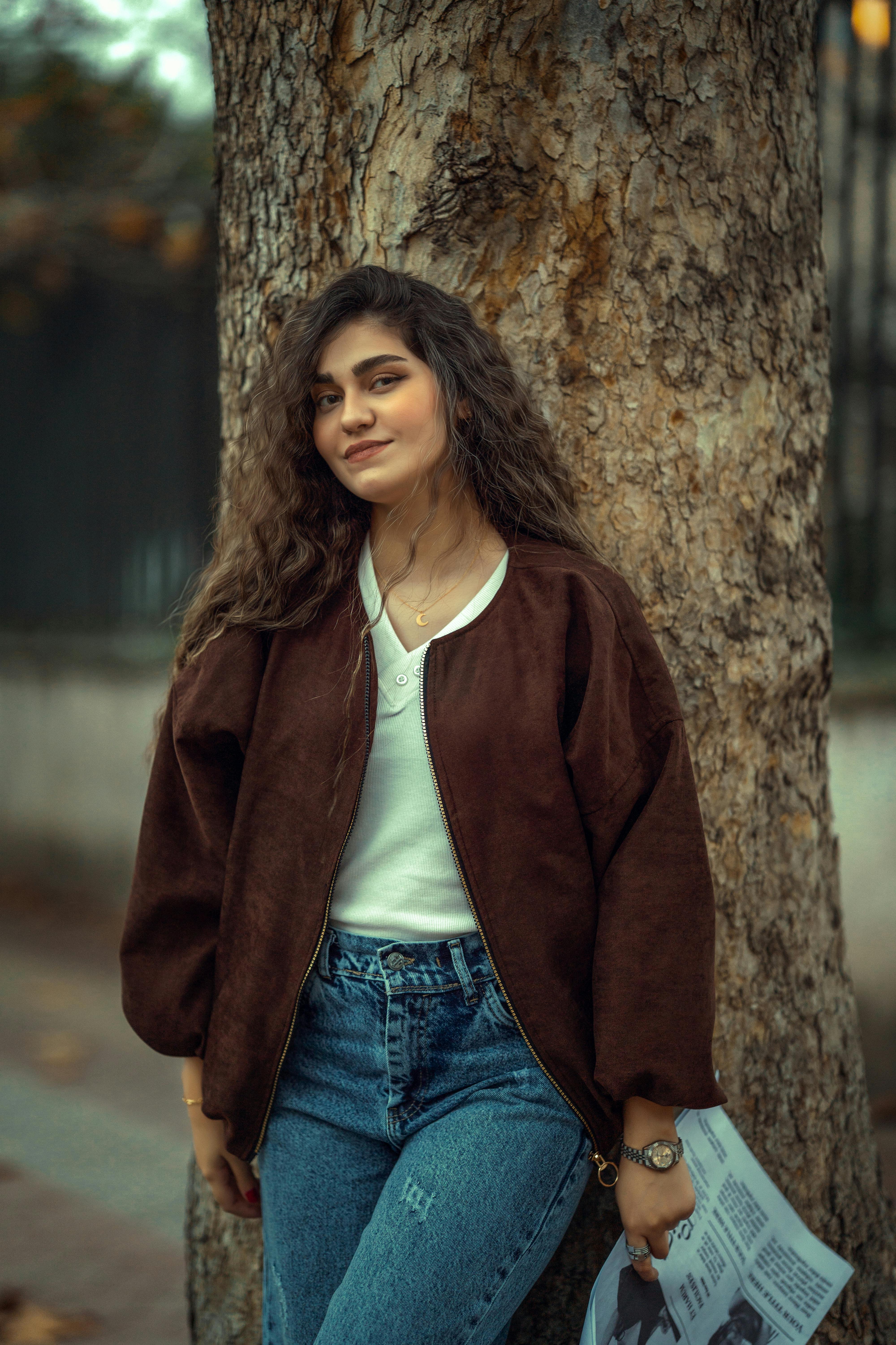 Stylish young woman with curly hair poses by a tree in fall attire, exuding confidence.
