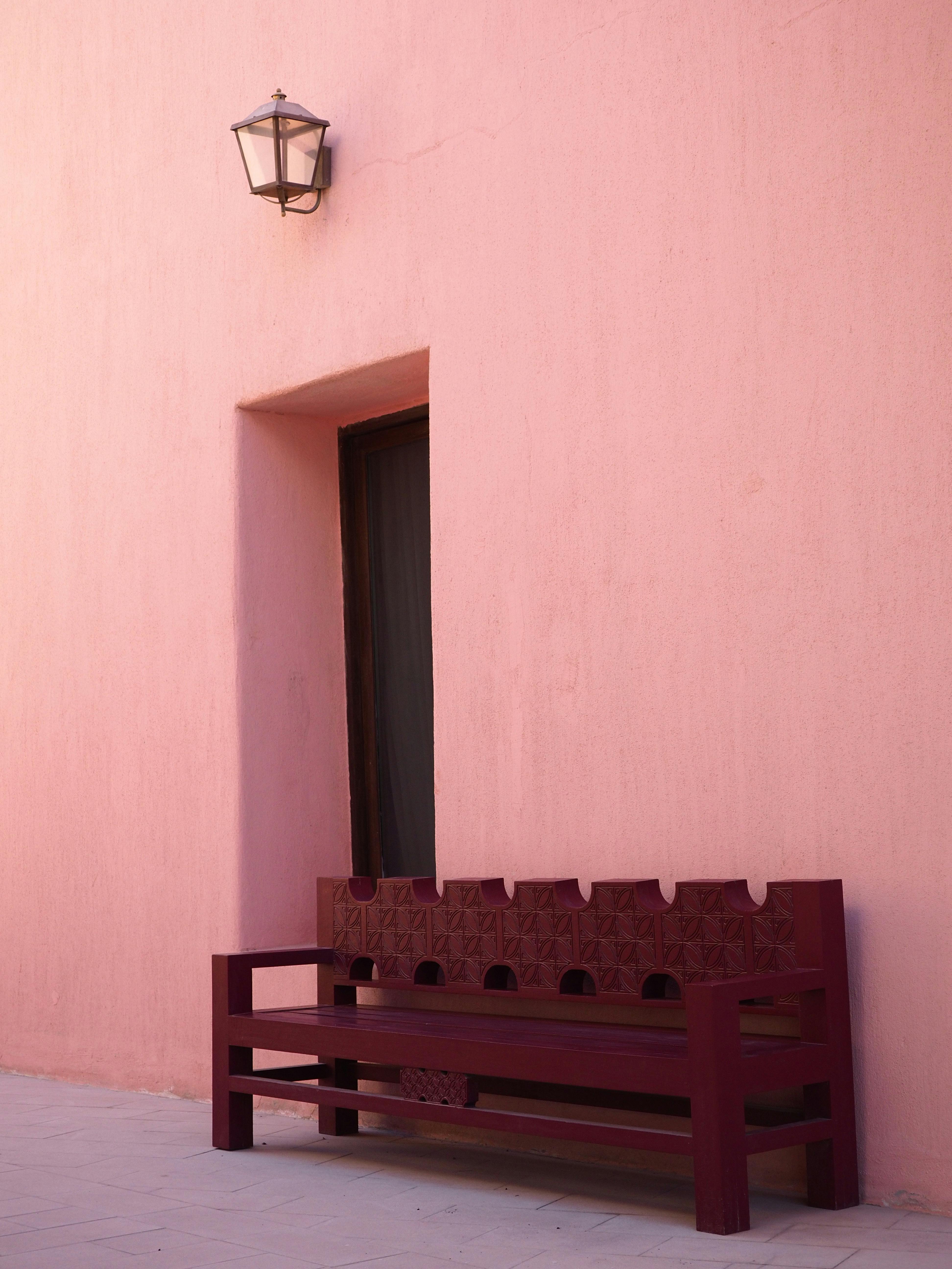 A minimalist outdoor scene featuring a pink wall, wooden bench, and lamp in Doha, Qatar.