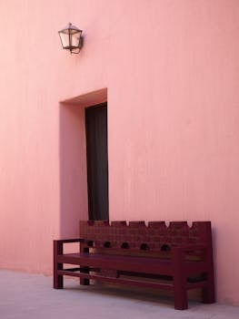 Solitude in soft light: a dark wooden bench rests beneath a vintage lantern on a smooth pink wall, exuding quiet elegance.         