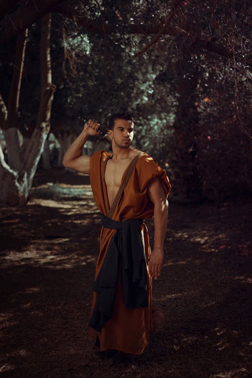 Man Wearing Orange Traditional Costume Holding Sword While Standing Surrounded With Green Trees
