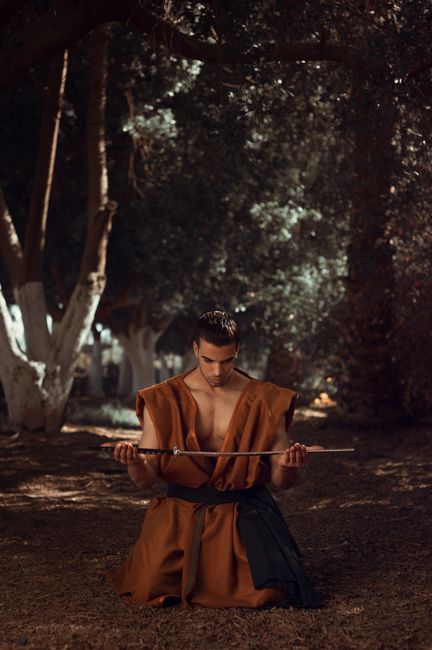 A young man kneeling with a sword in a wooded area, showcasing martial arts focus.