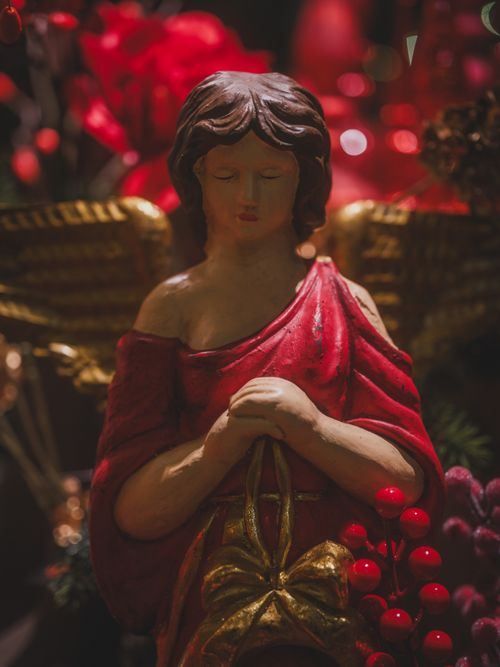 Free A serene angel sculpture with red robe surrounded by festive holly berries and warm lighting. Stock Photo