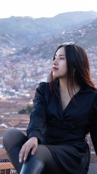 Stylish young woman in black sits overlooking a panoramic mountainous cityscape, exuding confidence.