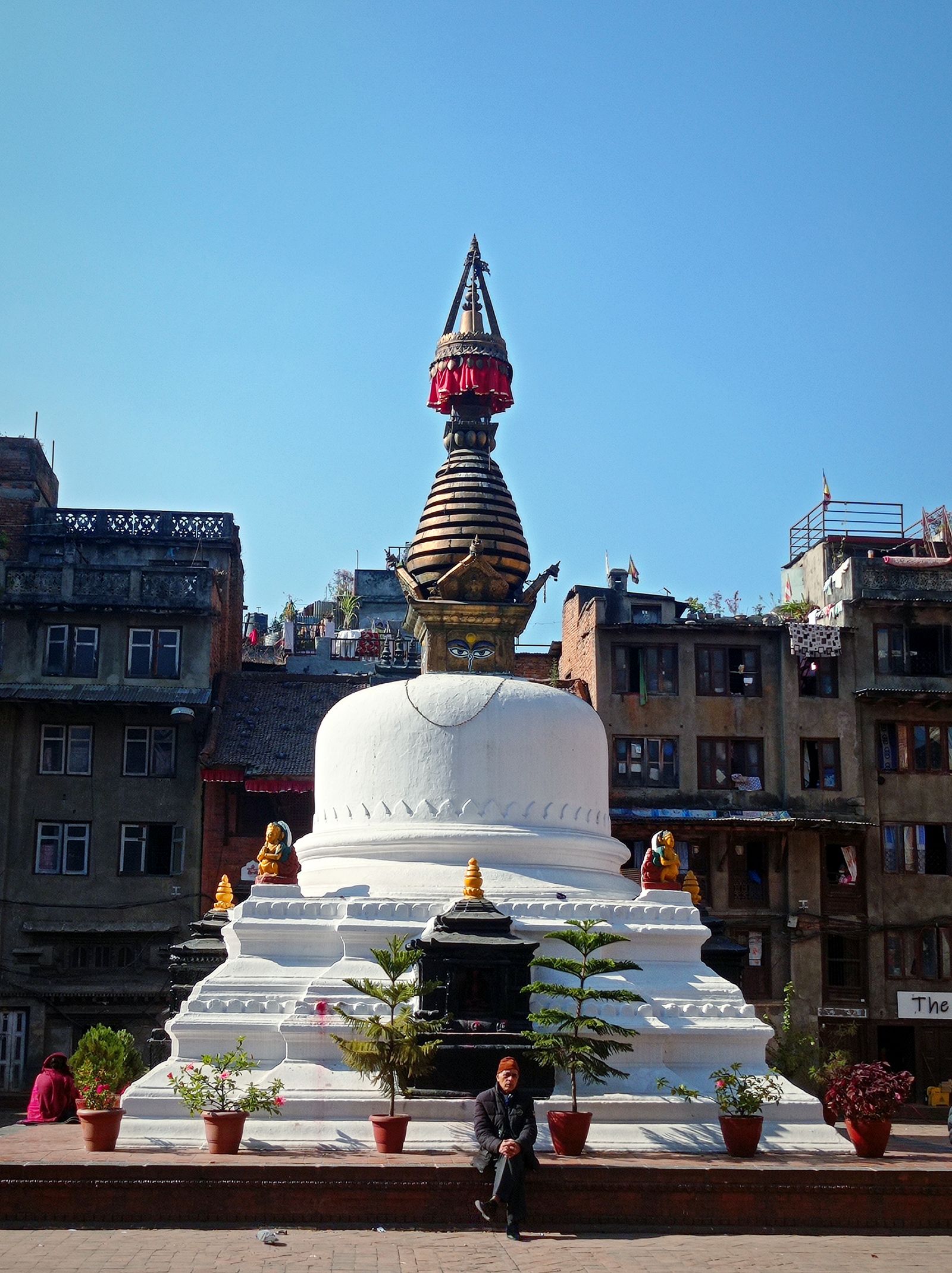 A small whitewashed stupa with a tiered spire in a square of old brick houses in Kathmandu, a man seated at its base