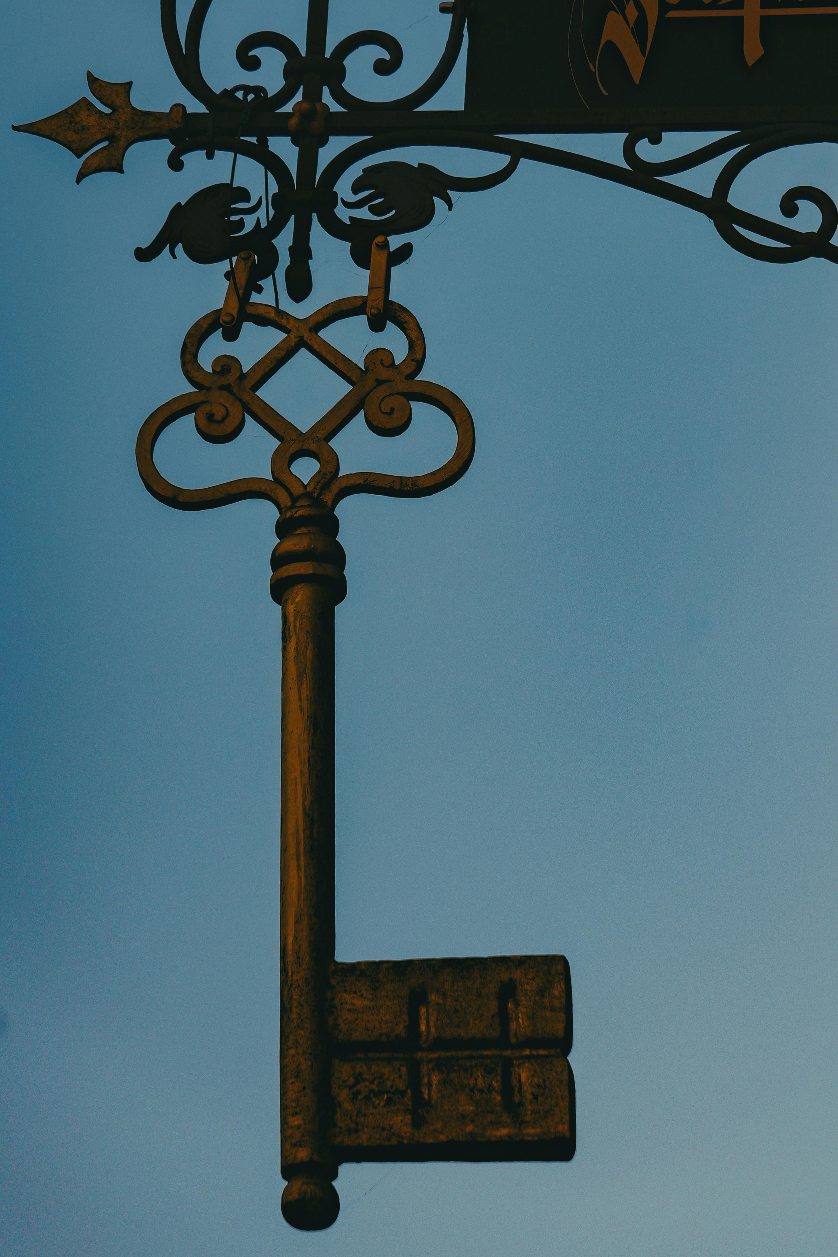Free An ornate vintage iron key sign hanging with a clear blue sky backdrop, perfect for decor and architecture themes. Stock Photo