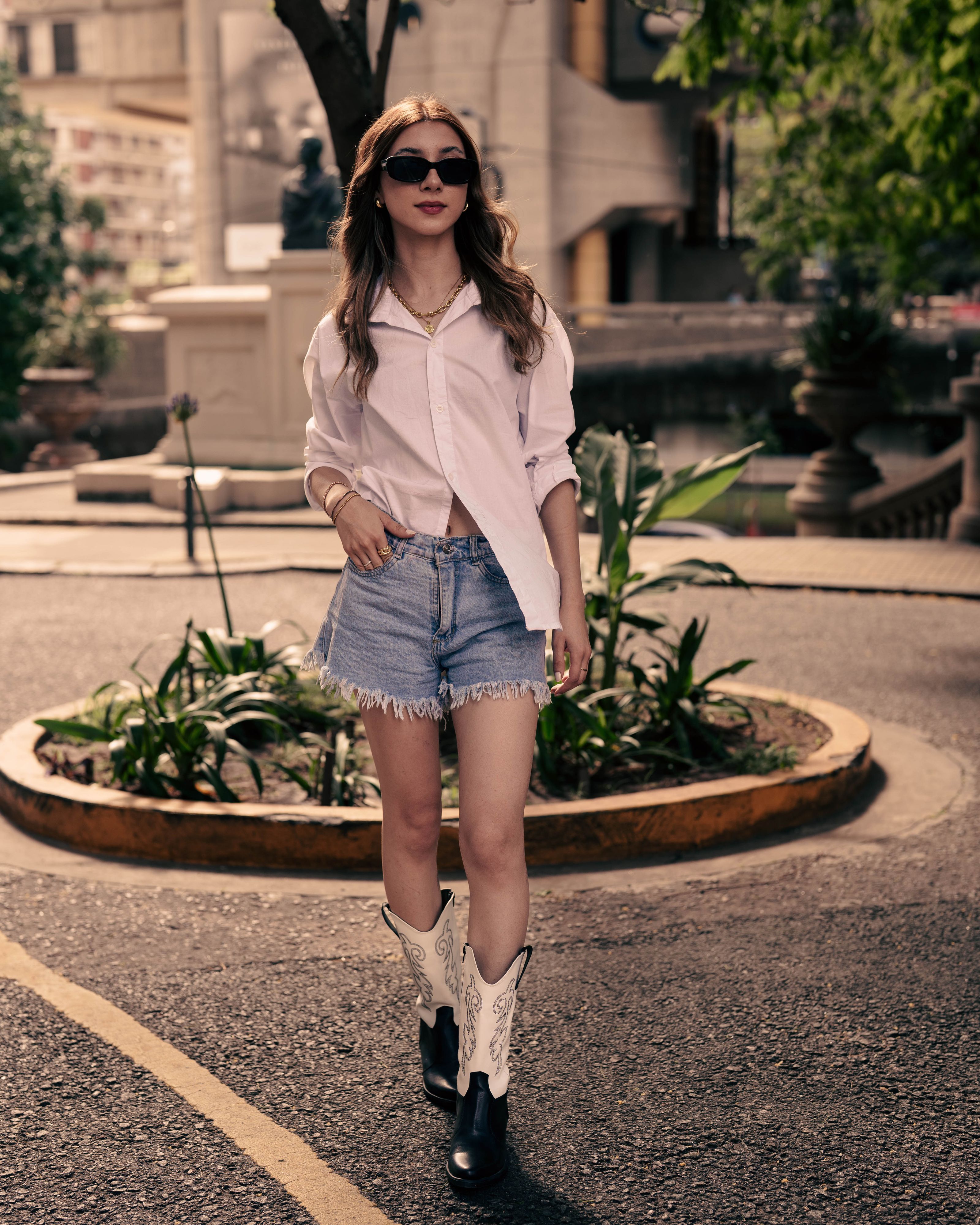 Mulher estilosa de shorts jeans e botas em uma cena urbana com vegetação e arquitetura moderna.