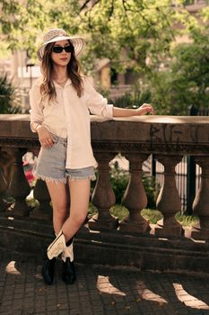 Fashionable woman in hat and sunglasses leaning on a stone railing in summer.