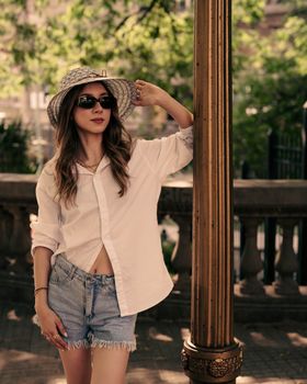 Stylish woman in sunglasses and hat, embracing summer fashion in an urban park.