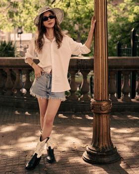 Fashionable woman in denim shorts and cowboy boots posing outdoors in summer light.