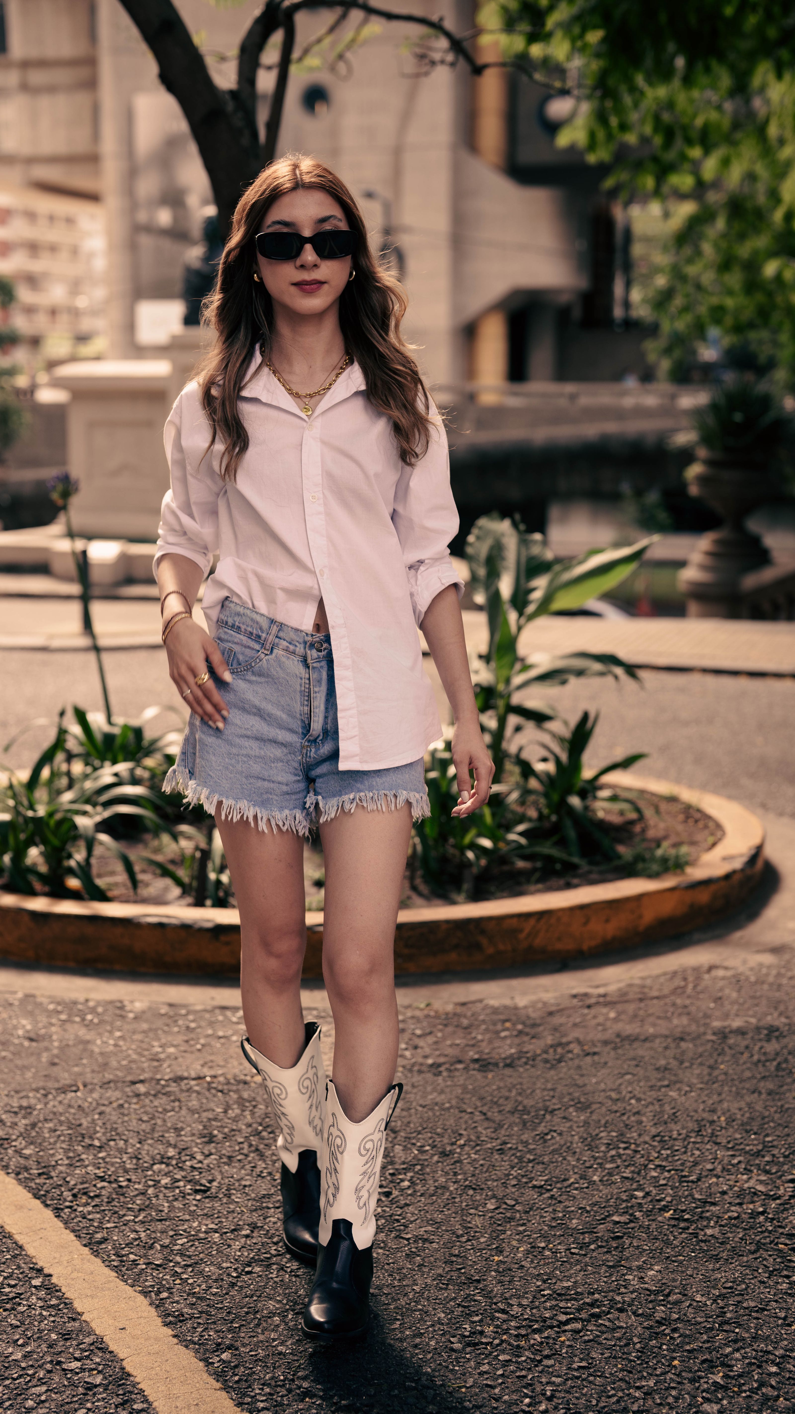 Mulher fashion em camisa branca e botas de cowboy caminhando ao ar livre.