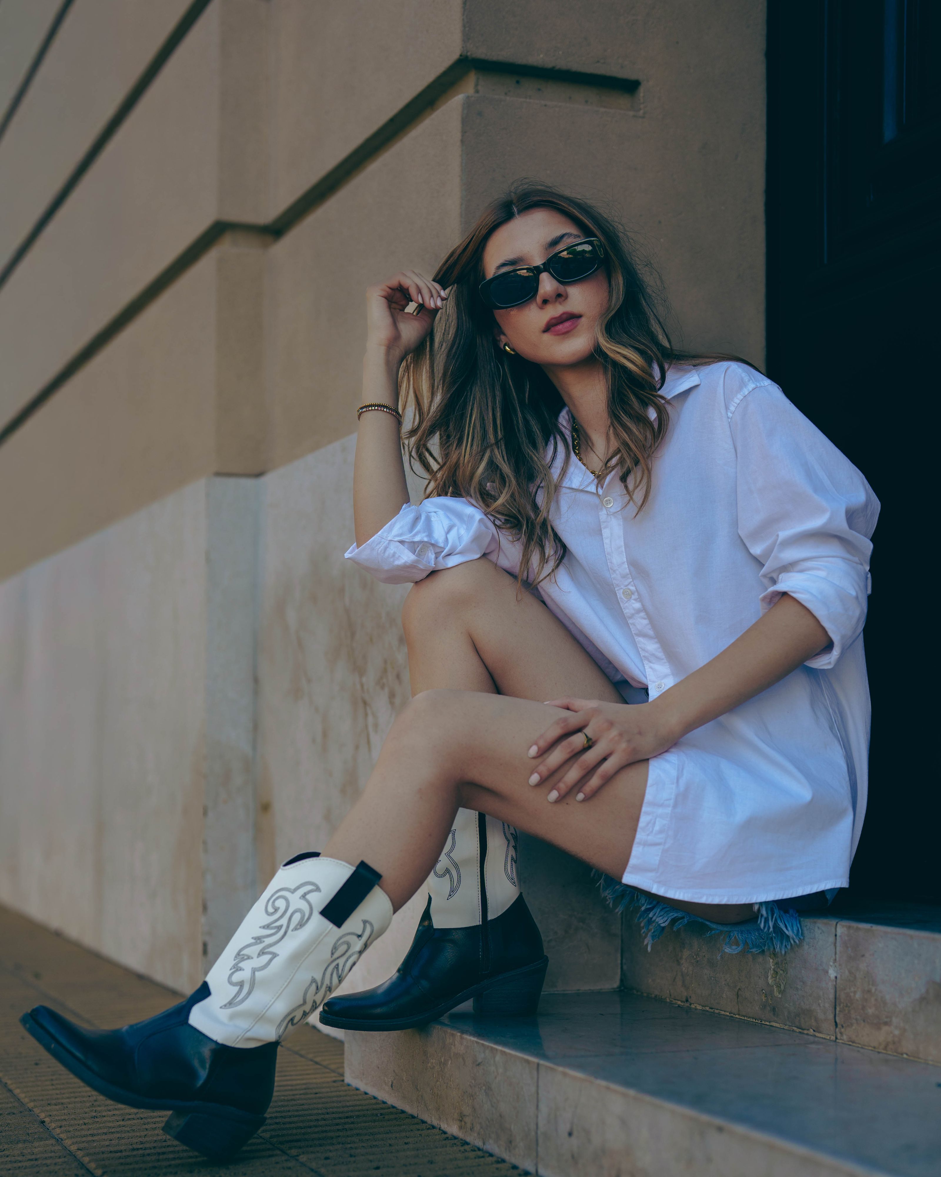 Mulher estilosa com uma camisa branca e botas de cowboy sentada na borda da calçada, exudando moda moderna.