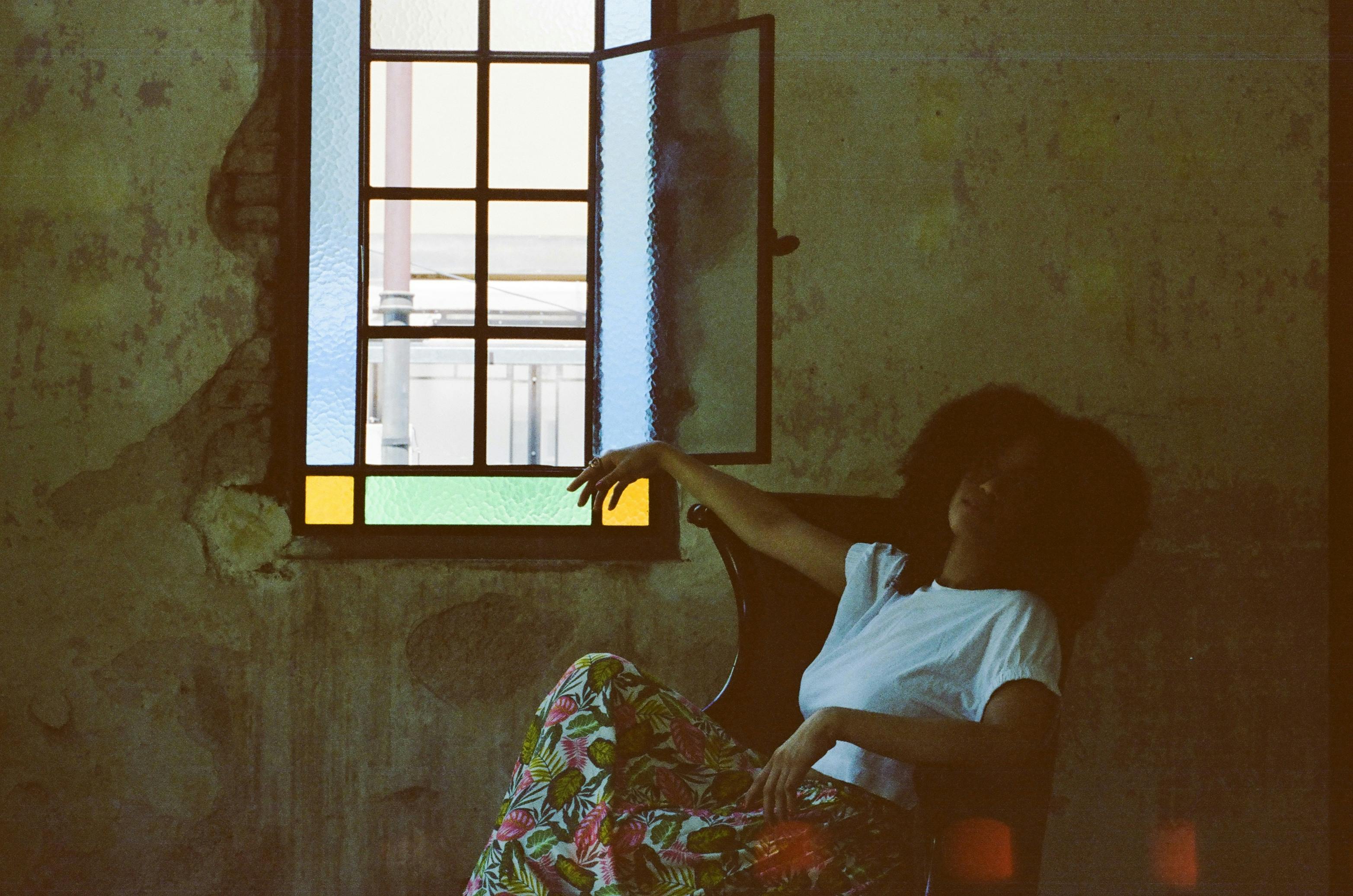 Kostnadsfria En kontemplativ kvinna sitter i ett svagt upplyst rum vid ett öppet glasmålningfönster och utstrålar en vintage- och konstnärlig stämning. Stock foto