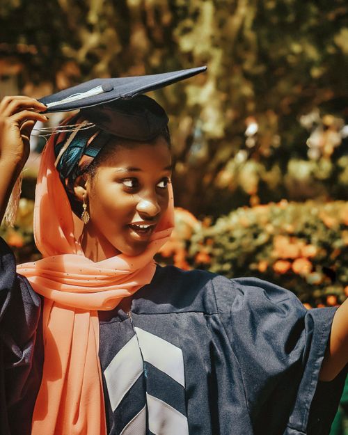 Free Happy young graduate adjusting her cap with a smile, celebrating her achievement outdoors on a sunny day. Stock Photo