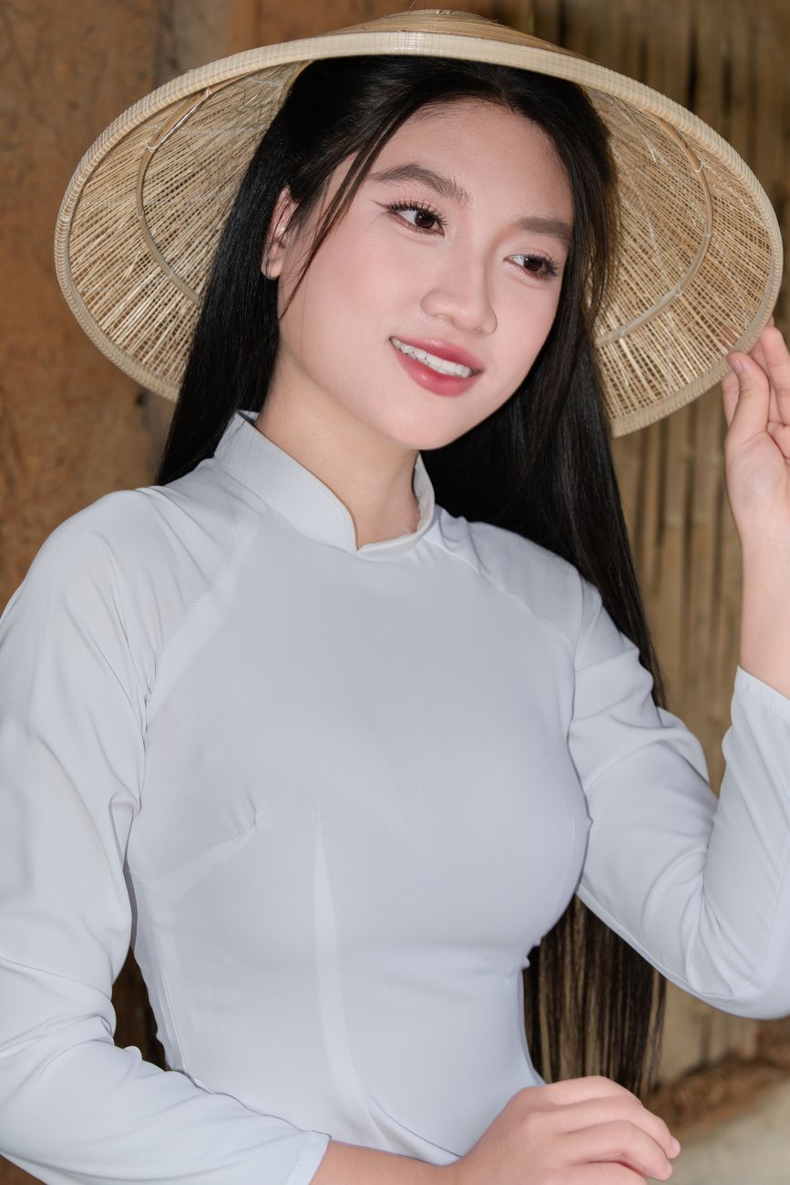 Asian woman in traditional attire and conical hat smiling confidently indoors.