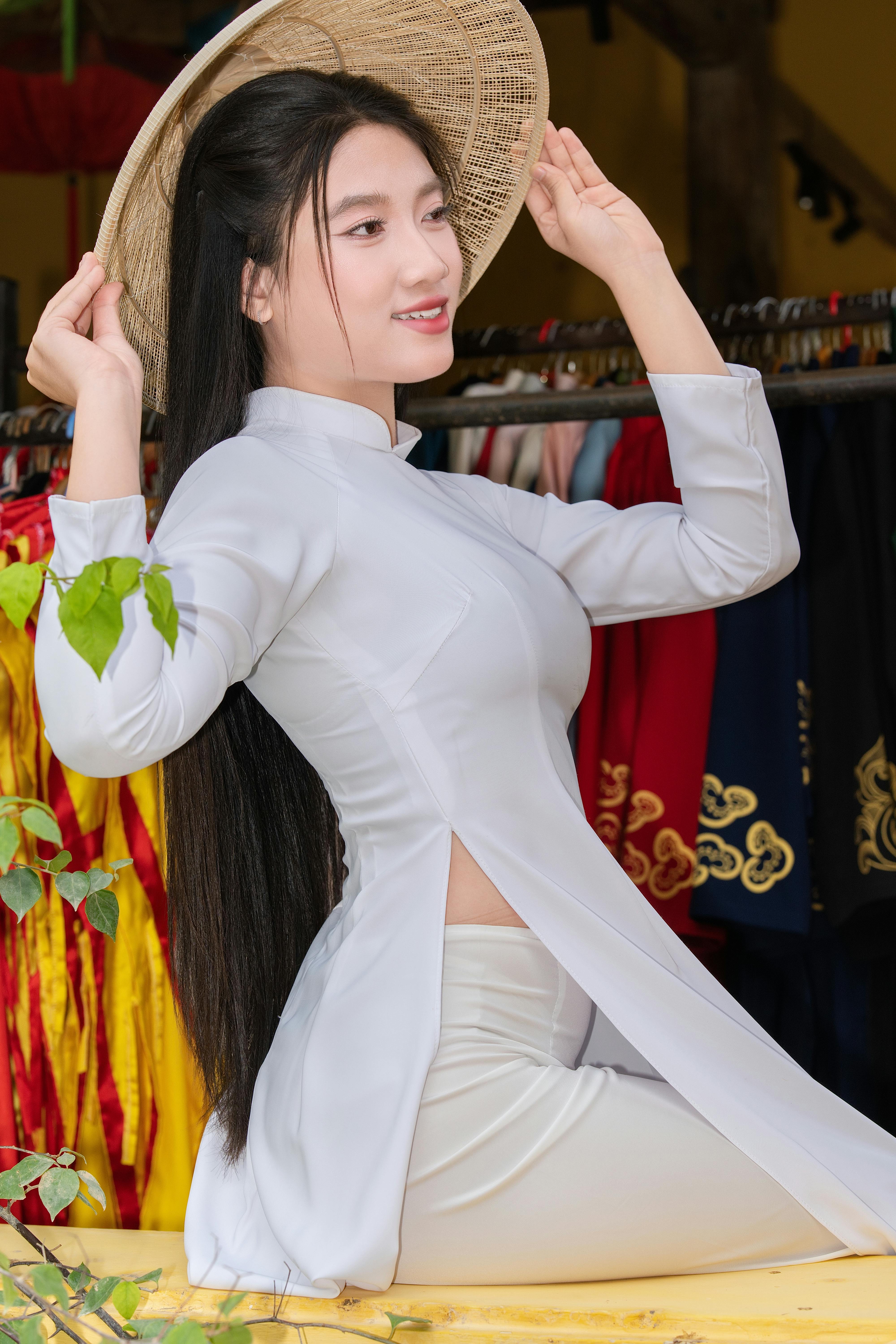Smiling woman in white ao dai adjusting a traditional conical hat indoors.