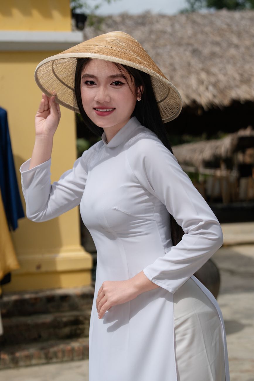 Woman in white ao dai and conical hat, smiling outdoors on a sunny day.