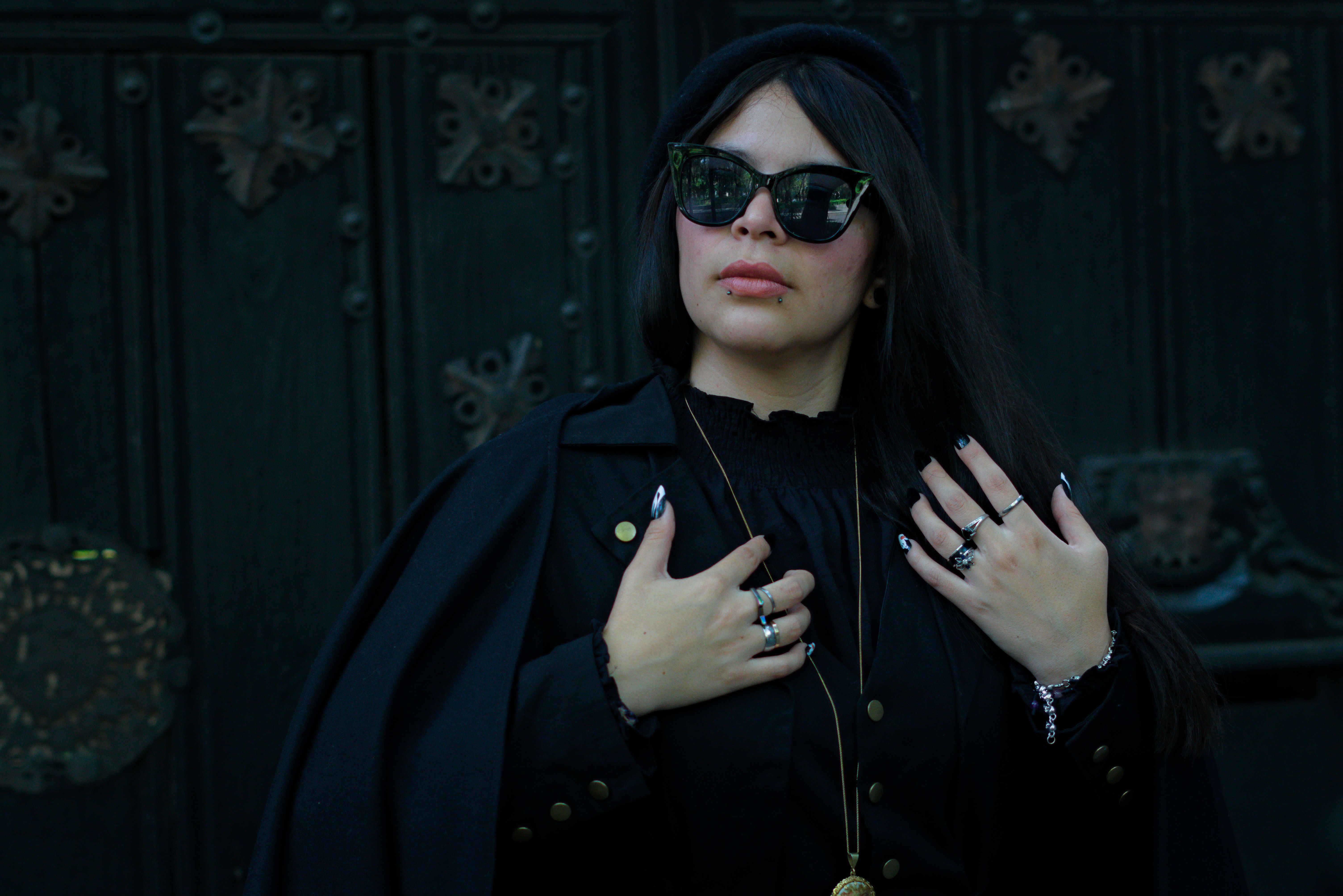 A fashionable woman in gothic attire poses confidently against a dark ornate background.