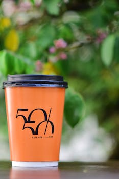 Vibrant orange coffee cup with black lid set against a lush green background outdoors.